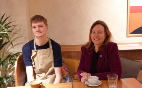 A young man in an apron sitting next to woman in a burgundy blazer drinking coffee