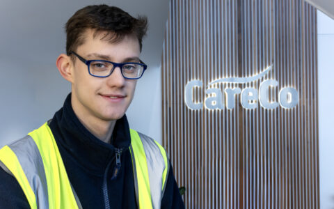 A young man in front of 'CareCo' sign wearing a high-ves