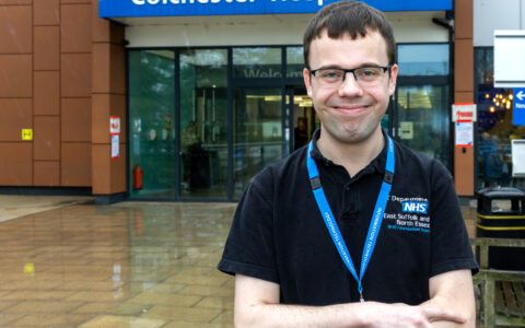 A young man standing in front of the Colchester Hospital looking at the camera and smiling