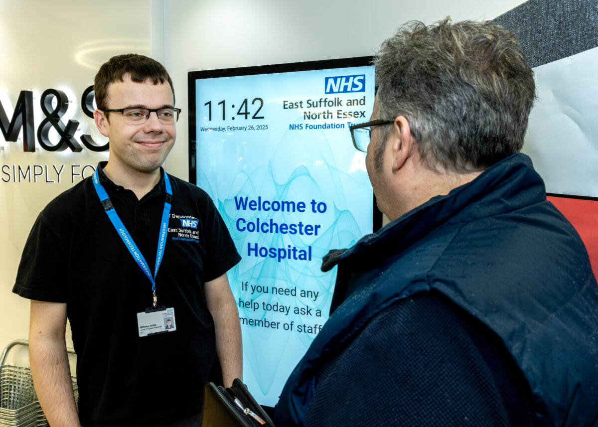 Two men talking to each other in front of a notice board saying 'Welcome to Colchester Hospital'