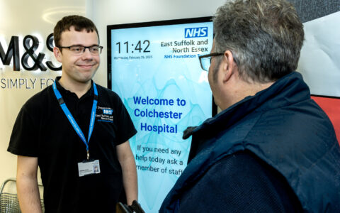 Two men talking to each other in front of a notice board saying 'Welcome to Colchester Hospital'