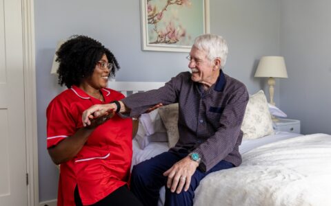 Woman in red tunic supports elder with rehabilitation near his bed-side