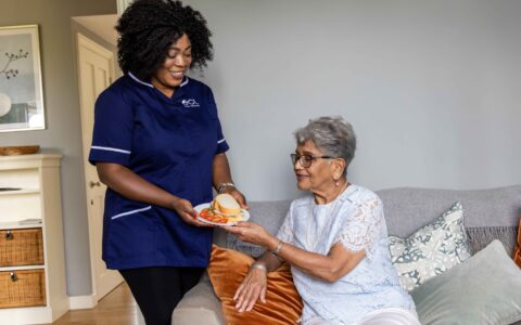 A woman in a blue tunic supporting and giving an item to an older woman who's sitting on a sofa.