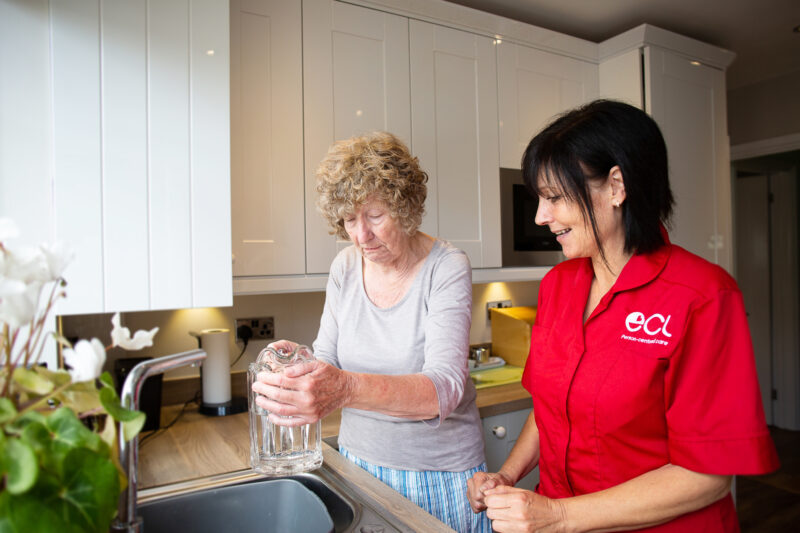 Older lady filling up a jug with tap water, supported by female care worker with black shoulder length hair and wearing a red ECL tunic.