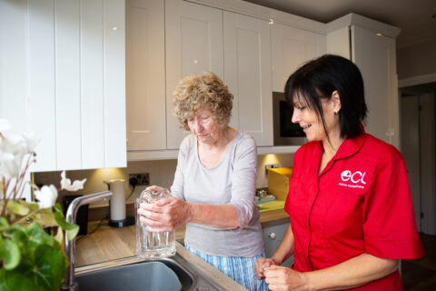 Older lady filling up a jug with tap water, supported by female care worker with black shoulder length hair and wearing a red ECL tunic.