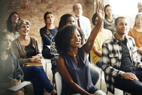 Group of people sitting down with a smiling female sitting at the front holding her hand up.