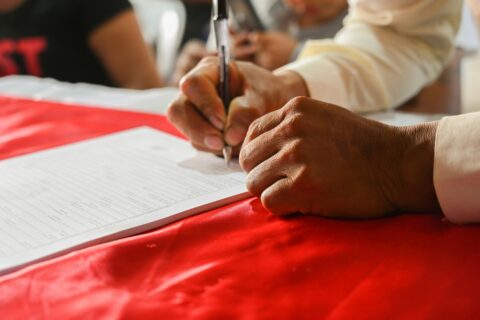 A man signing a form with his right hand and holding the document with his left. The table had a red runner on which the document sits.