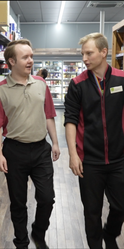 Young man with autism working at East of England co-op, talking to his colleague in the aisles.