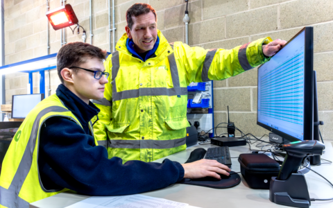 Two men in high-vis looking at a screen monitor, one of them is sitted, while the other is standing next to him and points at the monitor