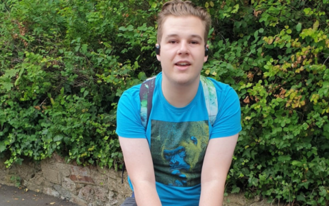 A blonde teenager wearing a blue tshirt with green graphic and a green backpack. He's posing on a statue of a hippopotamus.